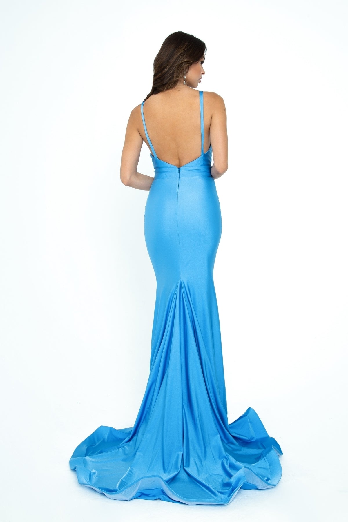 A woman poses against a white background, facing away to showcase the V-neck, backless design and thin straps of the bright blue Atria 6104H long prom dress by Atria Couture.